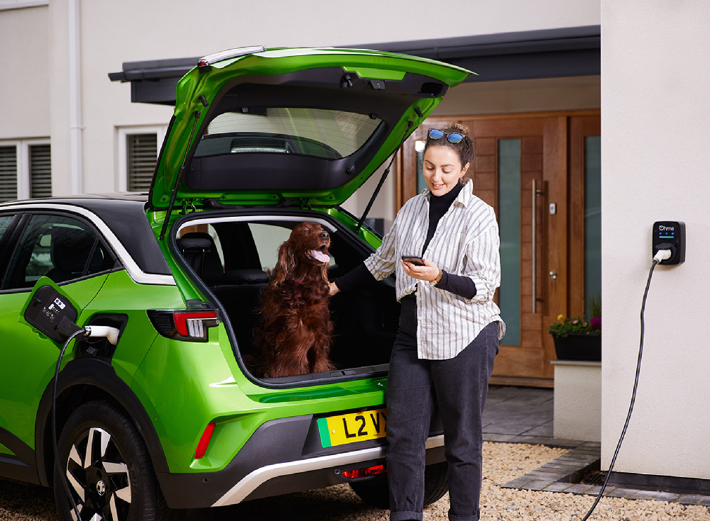 Woman using smartphone app while charging bright green Vauxhall Mokka electric vehicle with Ohme ePod EV charger, dog in boot